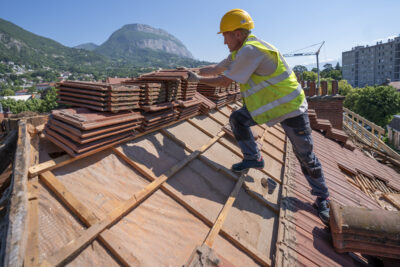 Un homme en casque et gilet jaunes de chantier, sur un toit, manipulant des tuiles - Agrandir l'image 3 sur 9, fenêtre modale
