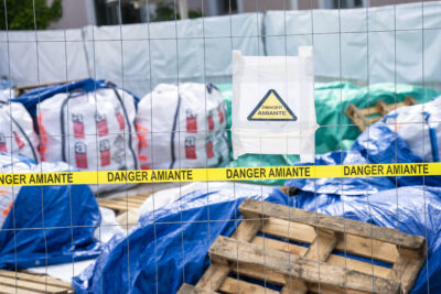 Un bandeau et une affiche Danger Amiante accrochés à un grillage derrière lequel se trouvent des gros sacs et des bâches - Agrandir l'image 2 sur 9, fenêtre modale