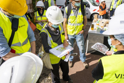 Un groupe de personnes portant des casques de sécurité blancs et jaunes, des gilets de chantier jaunes et des masques, examinant des documents, probablement des plans, lors d'une réunion en extérieur sur un site en travaux. - Agrandir l'image 1 sur 9, fenêtre modale