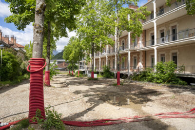 Une allée bordée d'arbres autour desquels sont enroulés des tuyaux rouges, avec en arrière-plan un bâtiment dont certaines fenêtres sont déposées. - Agrandir l'image 5 sur 9, fenêtre modale